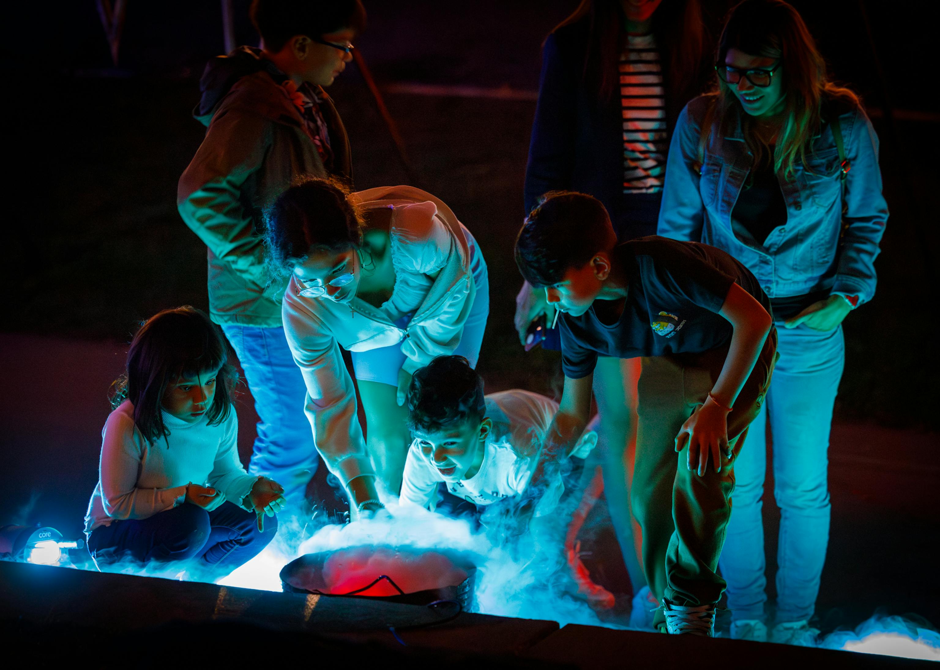 A group of young people looking down at a bucket filled with smoke which is lit in blue lighting