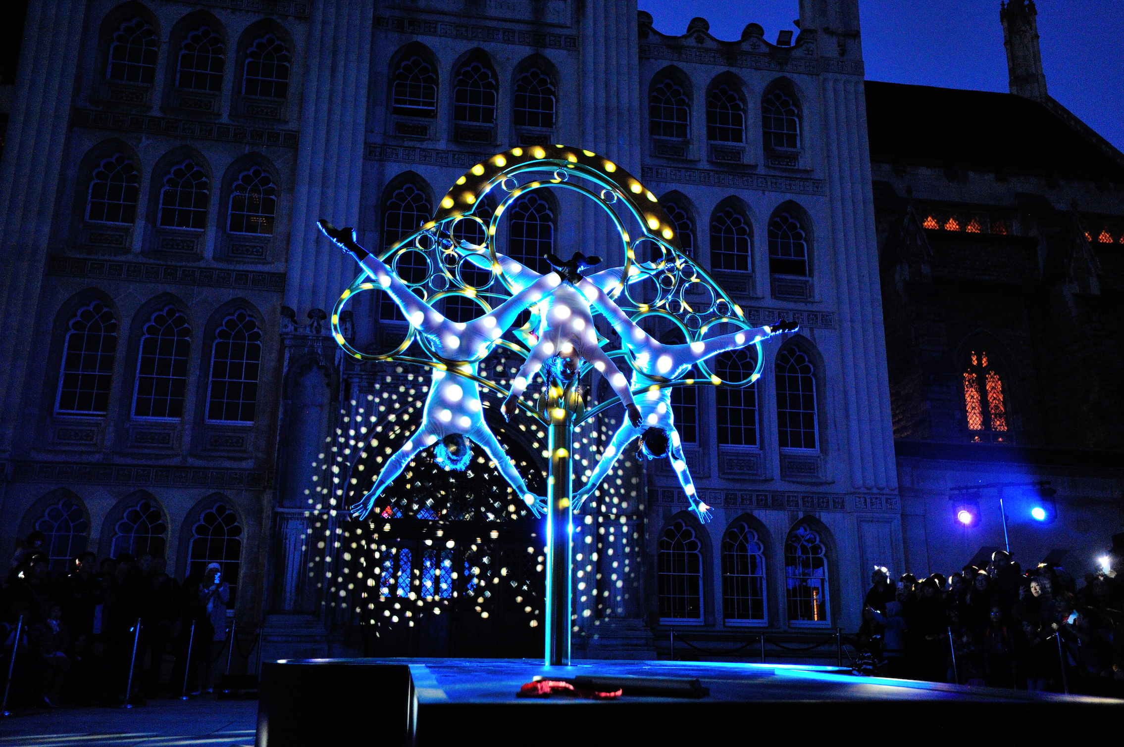 Three people hanging upside down from a large ornate key, with a projection of polkadots covering the people, the key and the building behind
