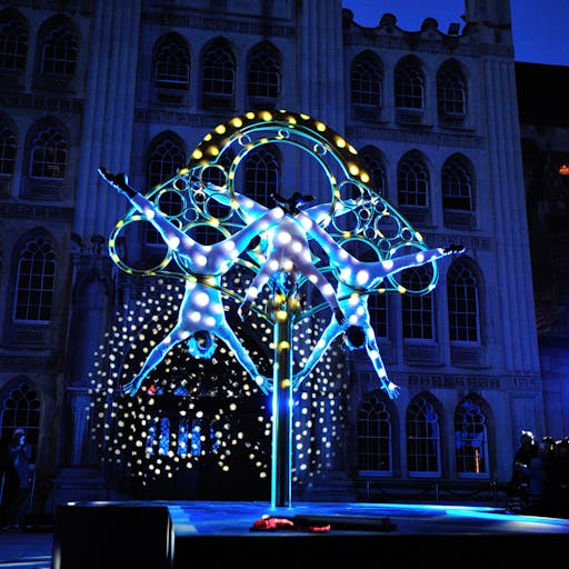Three acrobatic performers performing on a gaint scultpural golden key in the dark lit up by a project in front of an audience 