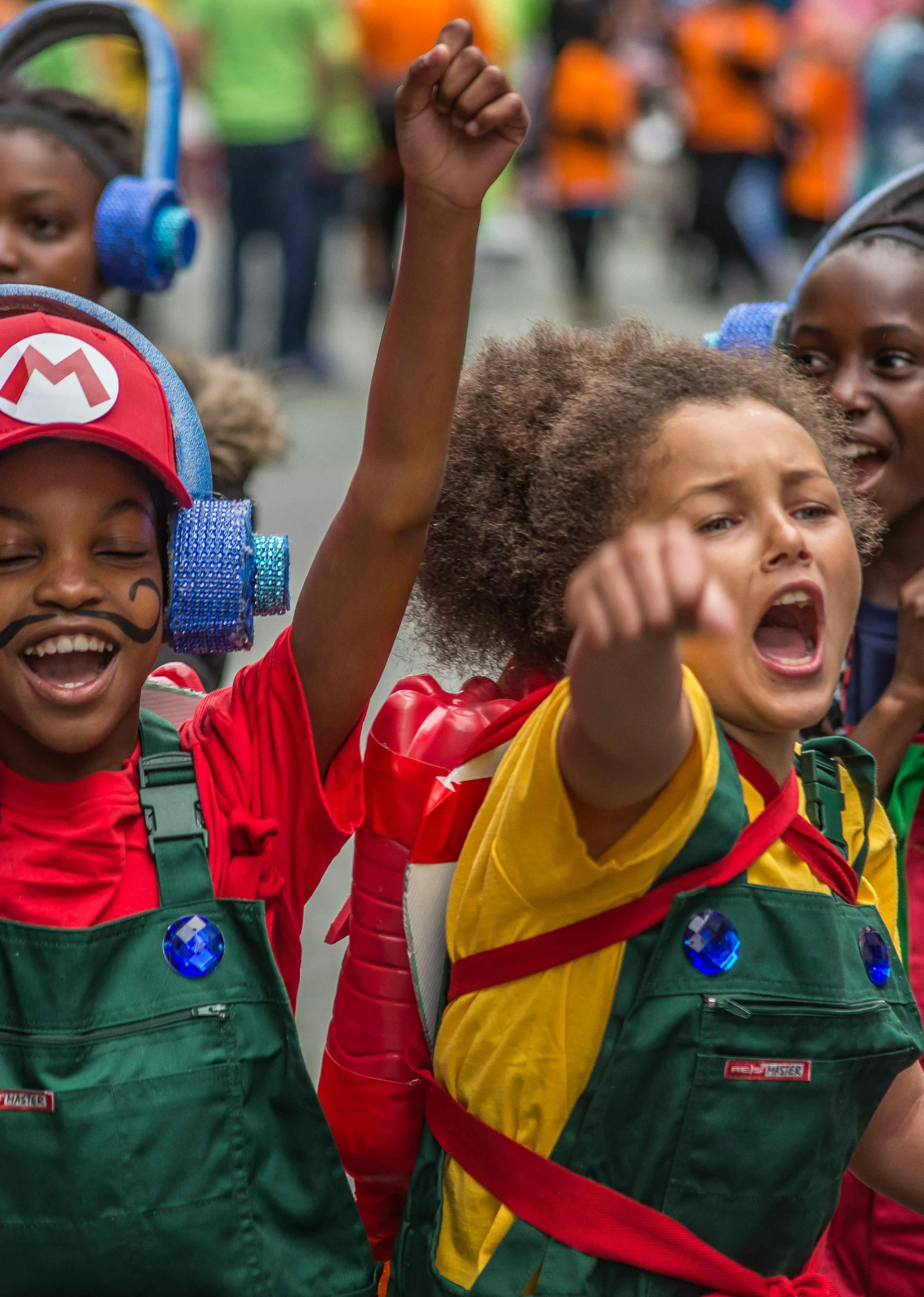 Two young people dressed up as Mario and Luigi with their hands up celebrating in front of a procession of people