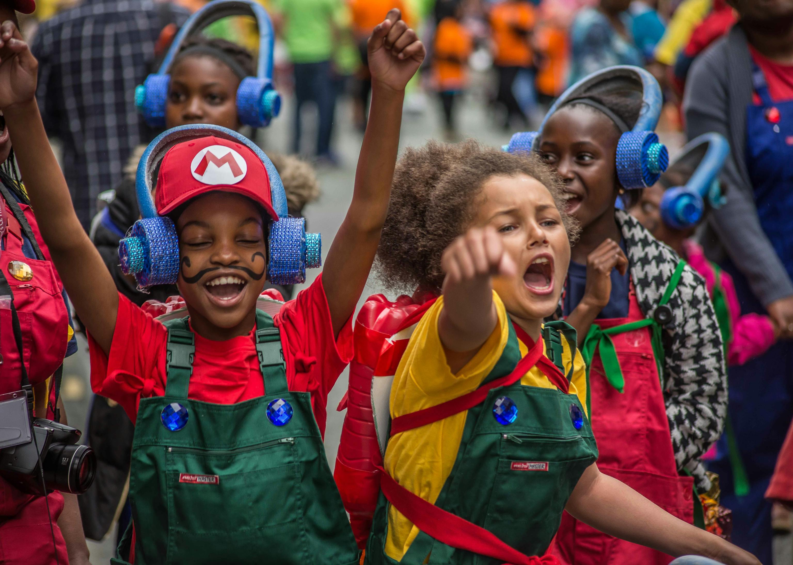 Two young people dressed up as Mario and Luigi with their hands up celebrating in front of a procession of people