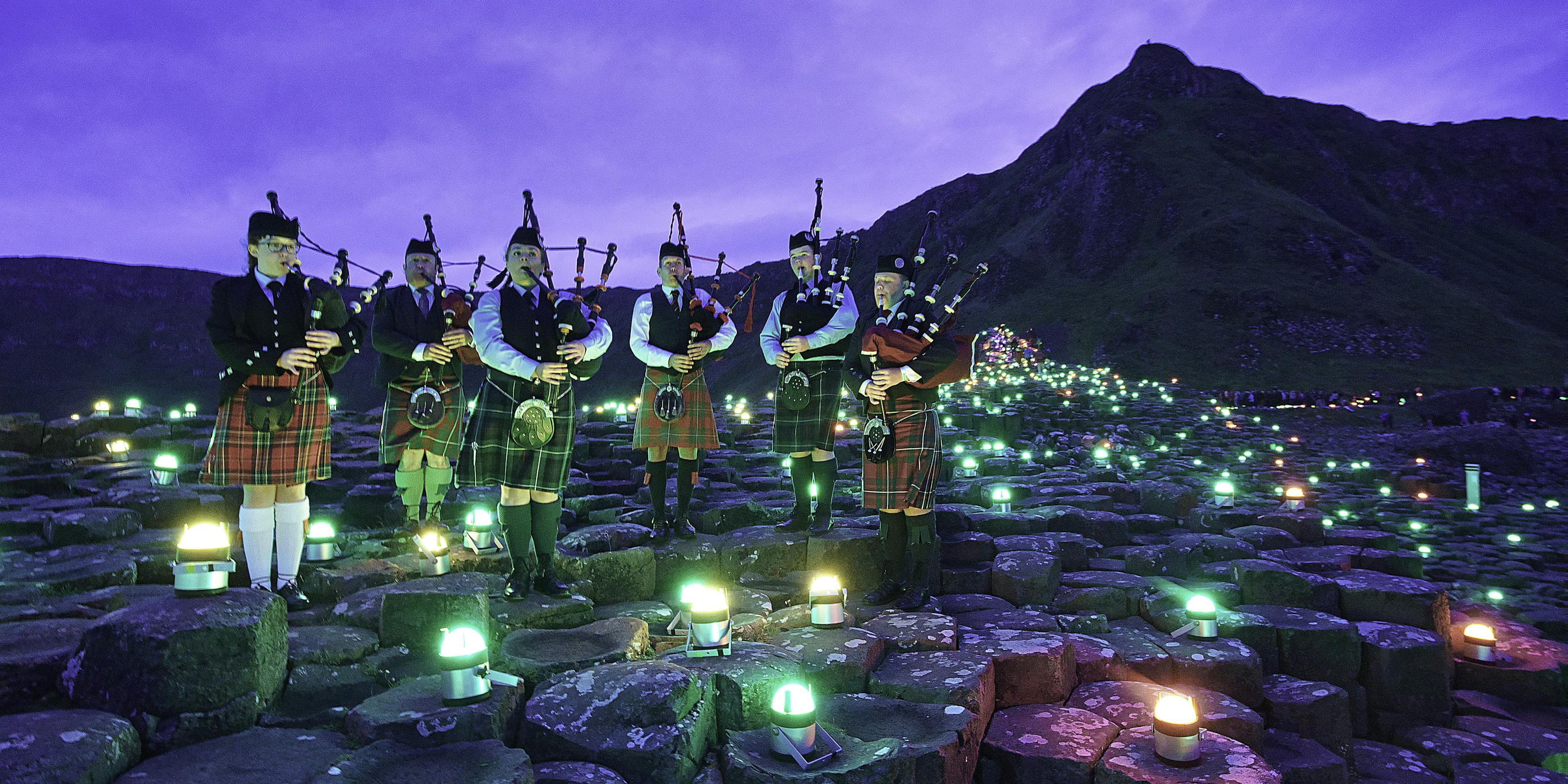 A group of six bagpipe players standing on the stones at Giant's Causeway surrounded by green Geolights