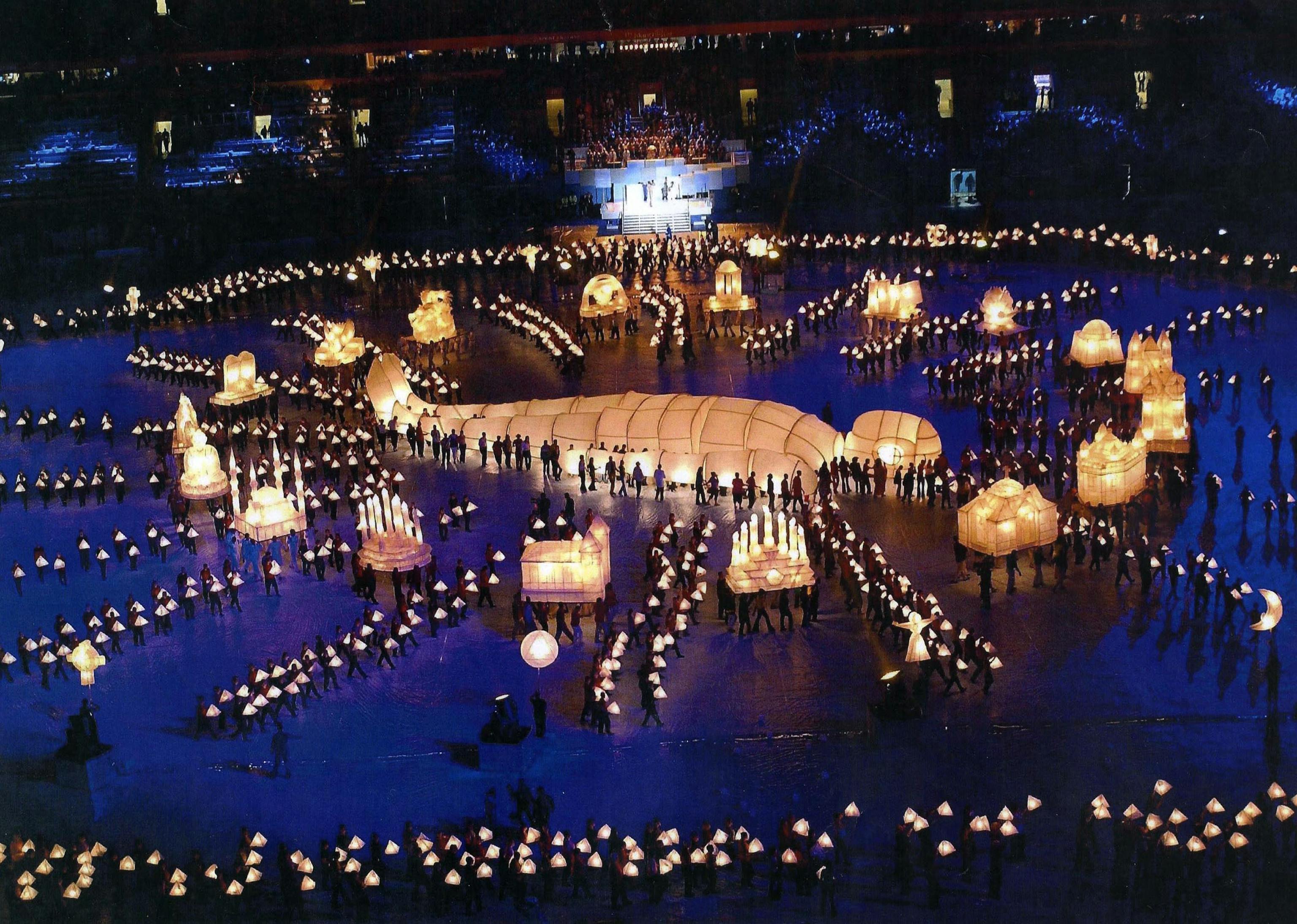 A large lantern shaped like a person lying in the centre of an arena and surrounded by hundreds of tiny dots of light, each a person holding a lantern