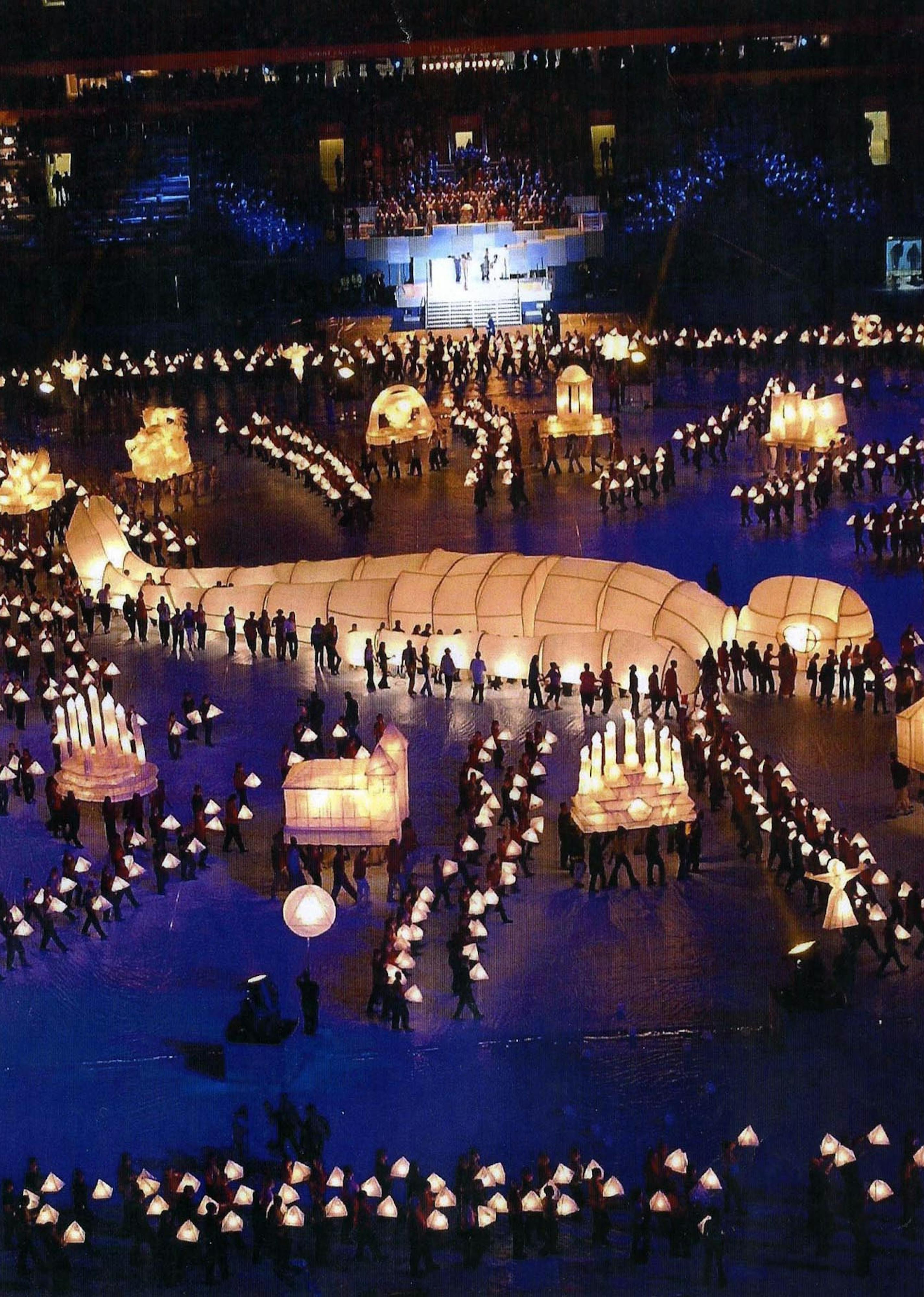A large lantern shaped like a person lying in the centre of an arena, it is surrounded by hundreds of tiny dots of light, each a person holding a lantern