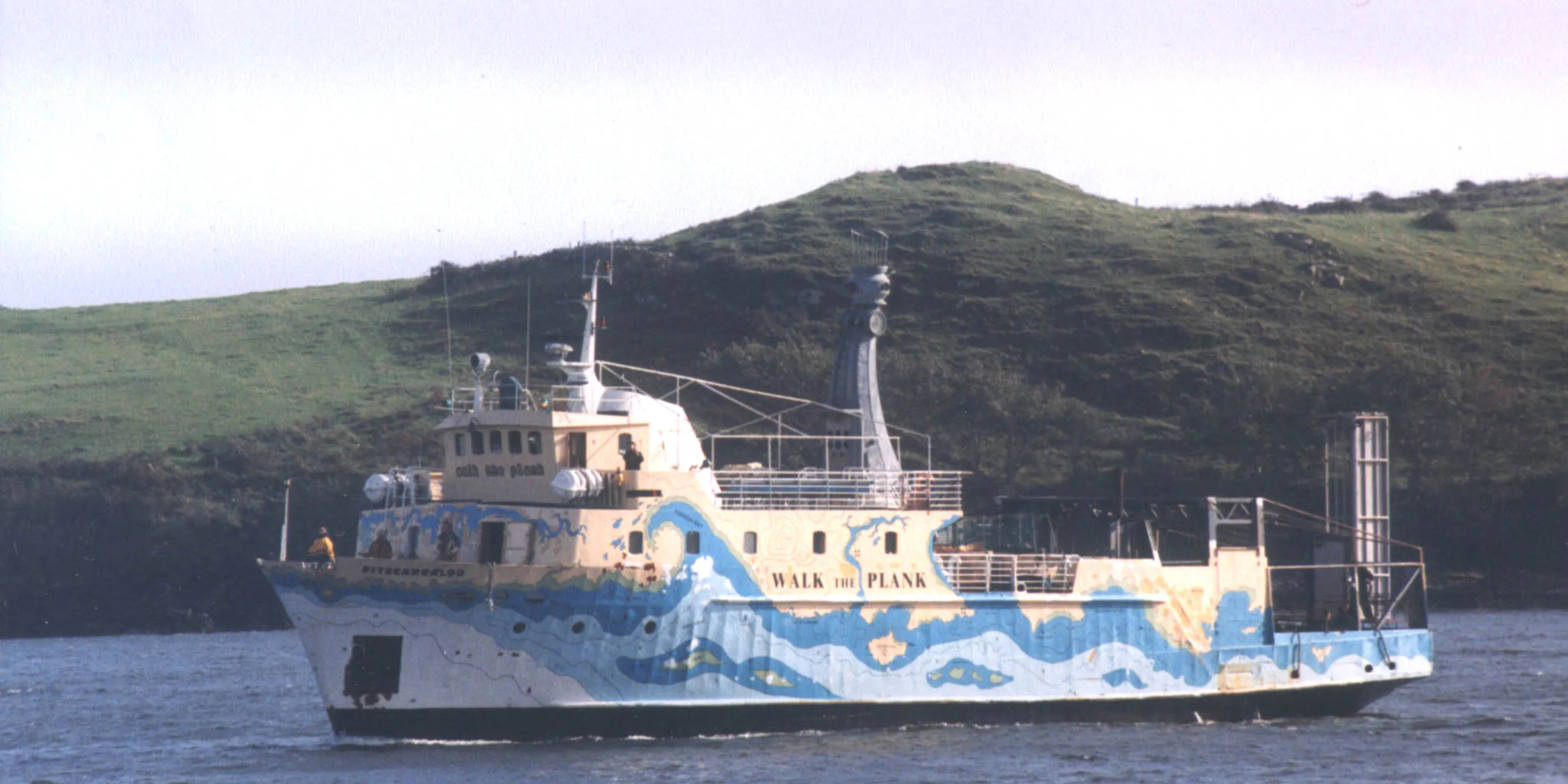 A blue and yellow ship that says Walk the Plank on the water in front of land