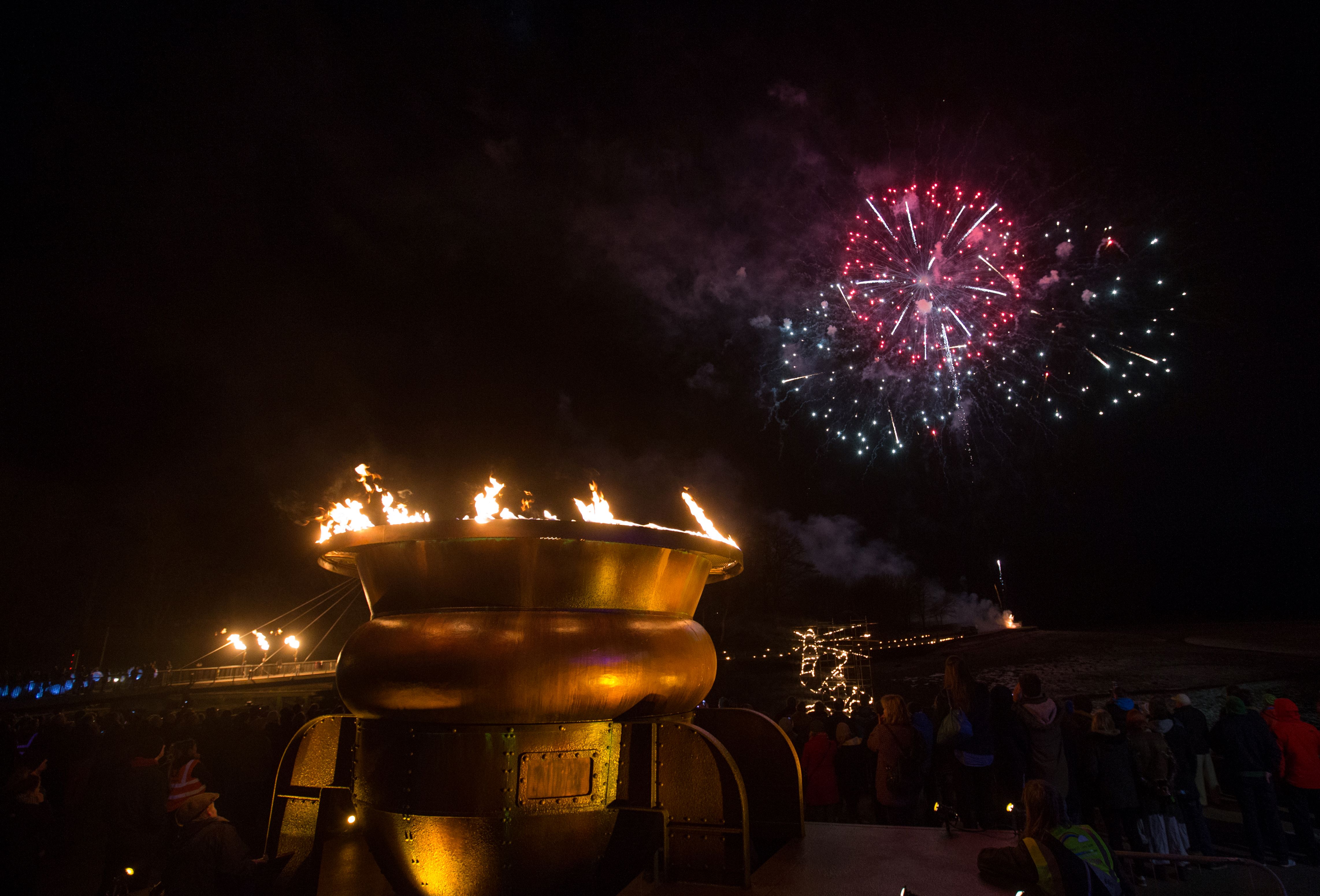 A large gold brazier with flames coming out of the top, against a night sky with fireworks