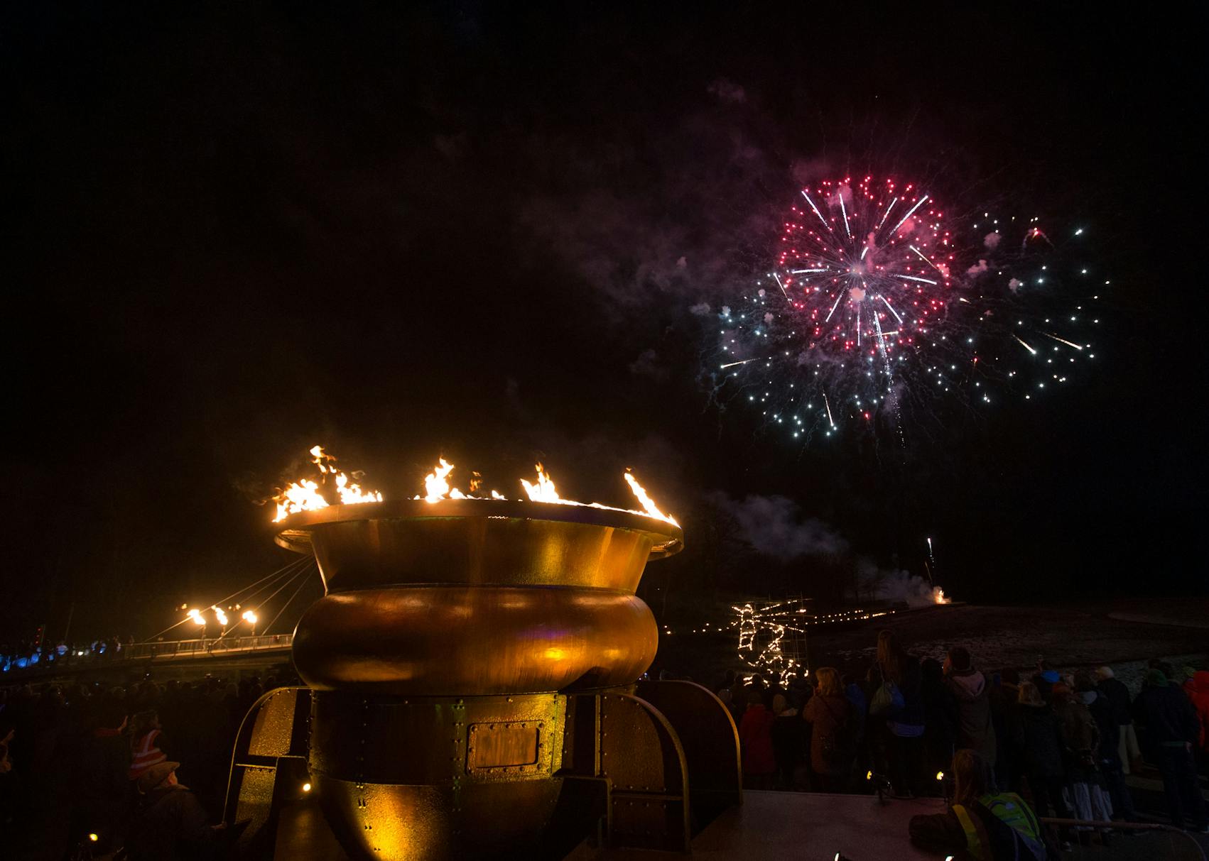 A large gold brazier with flames coming out of the top, against a night sky with fireworks
