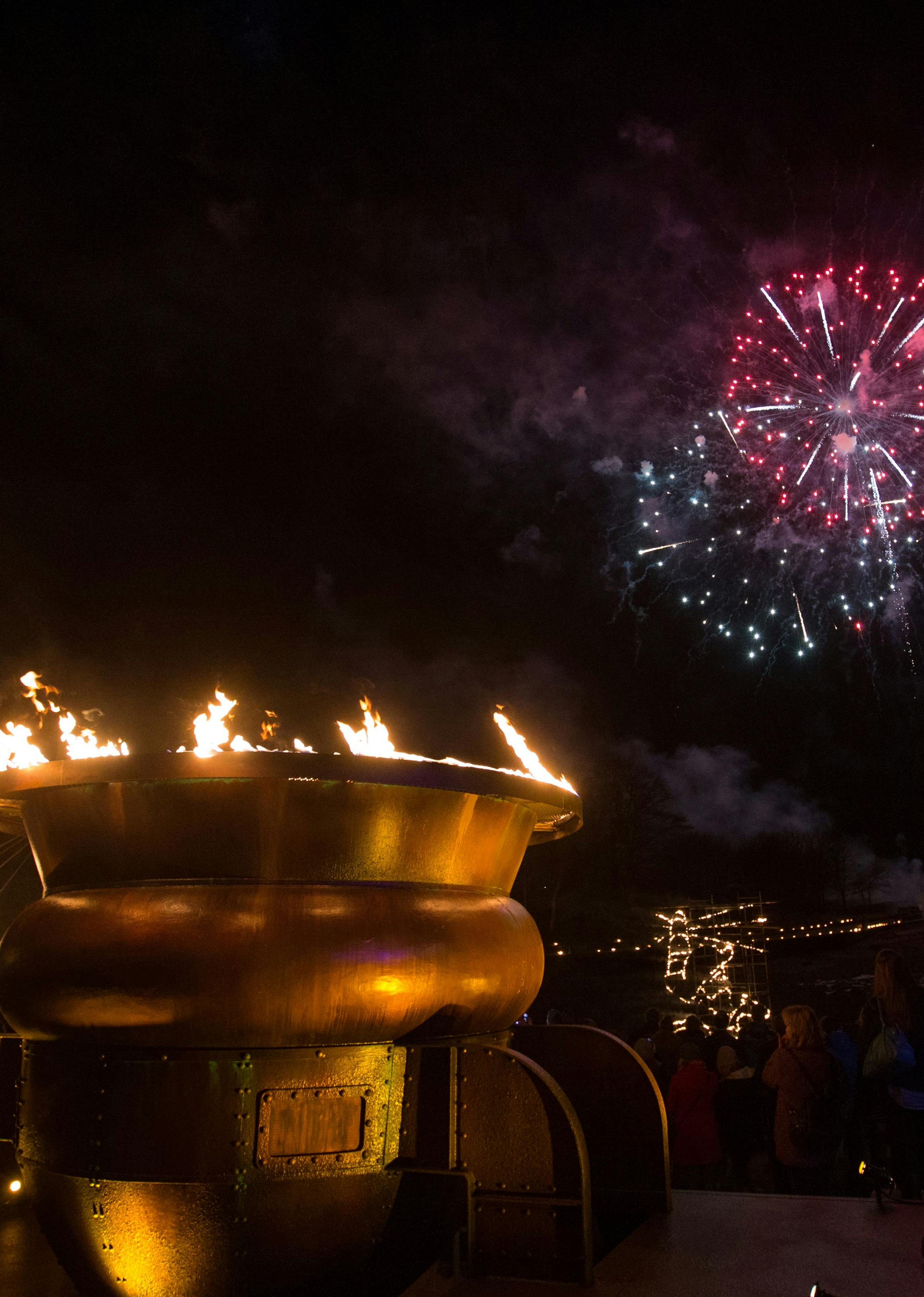 A large gold brazier with flames coming out of the top, against a night sky with fireworks