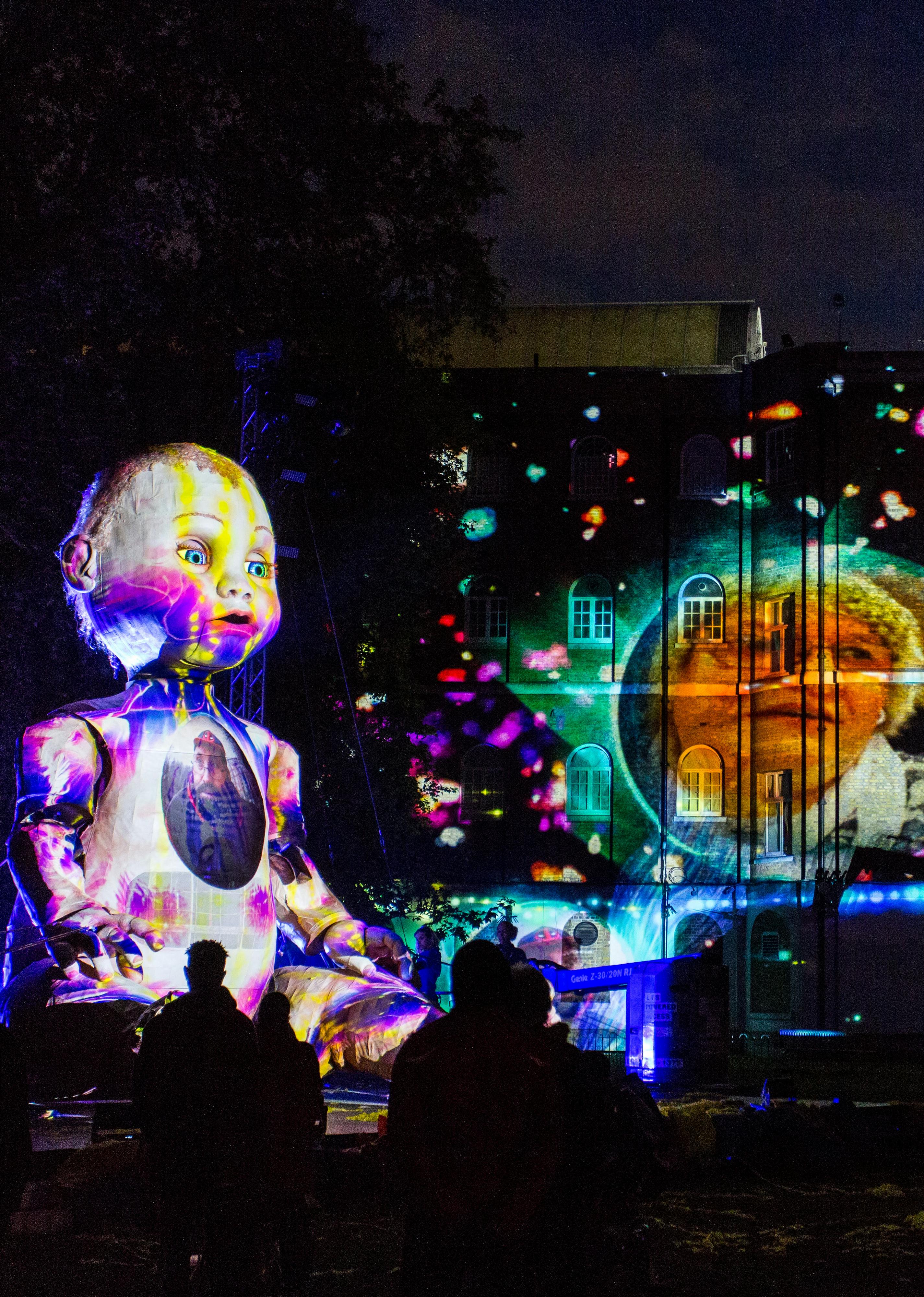 A giant puppet of a baby covered in projections that also cover the building next to it