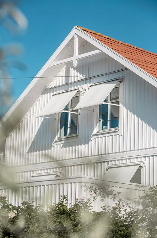 Två vita utvändiga fönstermarkiser är utdragna och skuggar två intilliggande övervåningsfönster på en vit träfasad av ett hus med rött tegeltak.