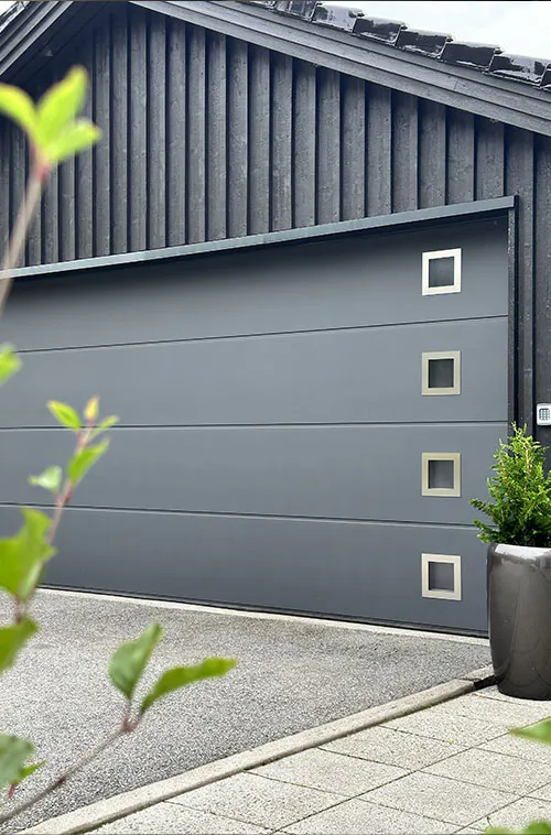 En slät mörkgrå garageport står stängd mot en svart träfasad; fyra kvadratiska fönster i högerkant, asfaltuppfart, krukväxt vid sidan och suddig grönska i förgrunden.
