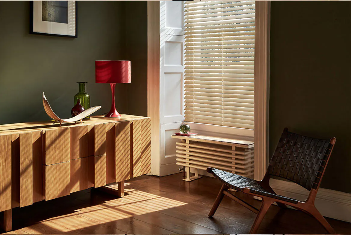 Persienner filtrerar mjukt dagsljus och kastar ränder av ljus över trägolv och sideboard i ett varmt vardagsrum med röd bordslampa, liten sittbänk och rottingstol.