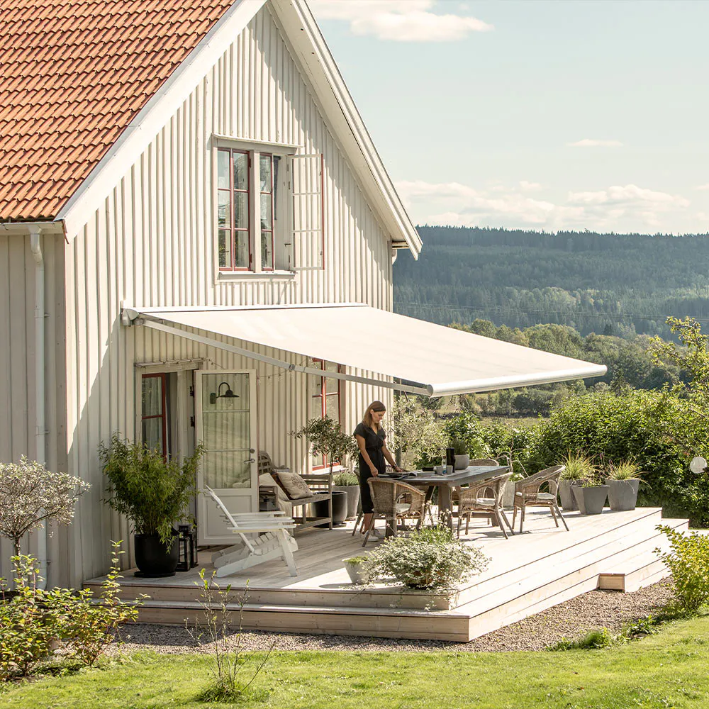 Markis utfälld över träaltan; en kvinna dukar ett bord på verandan; vitt trähus med rött tak, krukväxter, gräsmatta och vidsträckt skogssjö i bakgrunden.