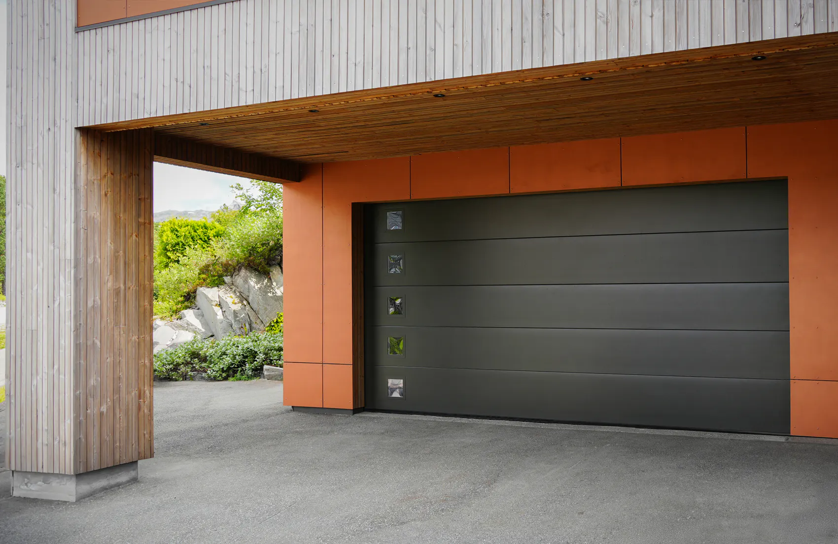 Stor mörkgrå garageport (sektionerad) står stängd i en modern fasad klädd i trä och orange paneler; med fyra små rutformade fönster, uppfart och gröna buskar till vänster.