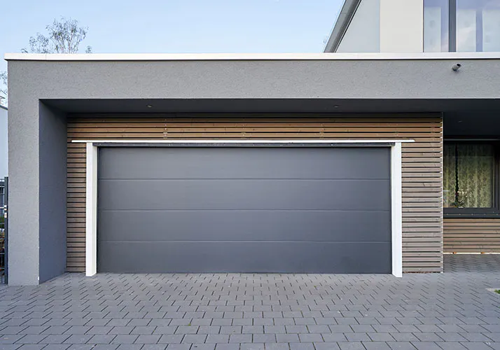En grå sektionerad garageport står stängd inom en modern fasad; vita karmar, horisontella träpaneler runt öppningen och stenlagd uppfart i förgrunden under blå himmel.