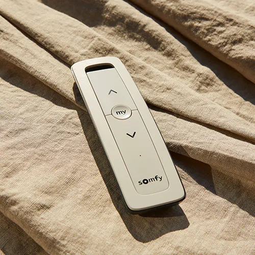 Somfy-fjärrkontroll vilar på skrynkligt, sandfärgat linne i mjukt solljus; den ligger stilla med upp- och nedpilar samt en "my"-knapp. Text: "my", "somfy", pilar "?" "?".