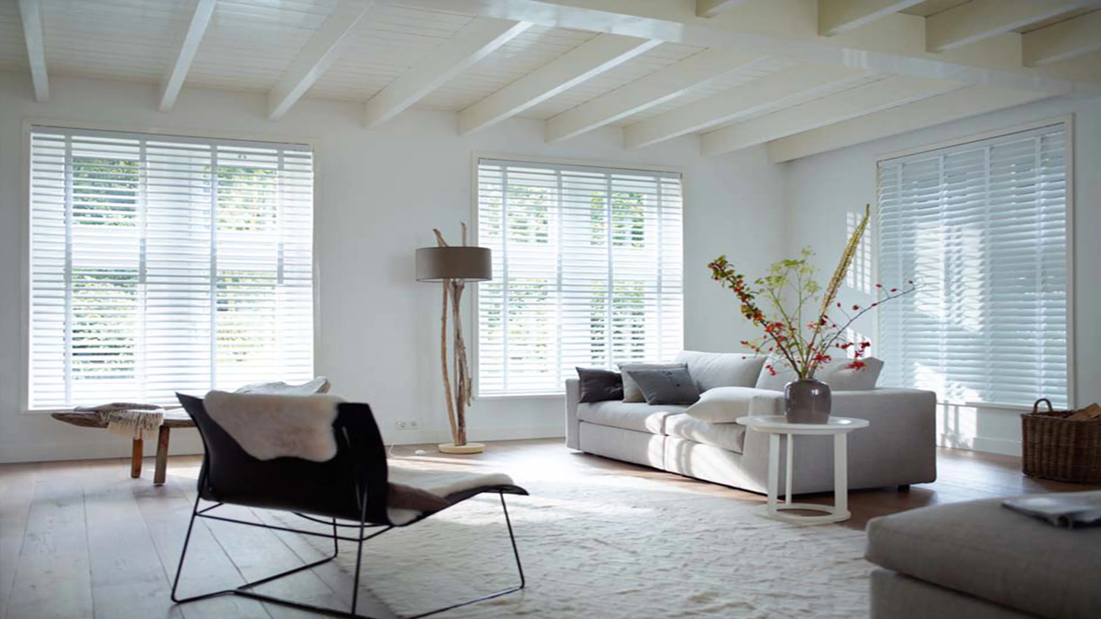 Vita jalusier för fönster släpper in diffust dagsljus i ett ljust vardagsrum med vit soffa, litet runt bord med vas, modern fåtölj, golvlampa och vitt träbjälktak.
