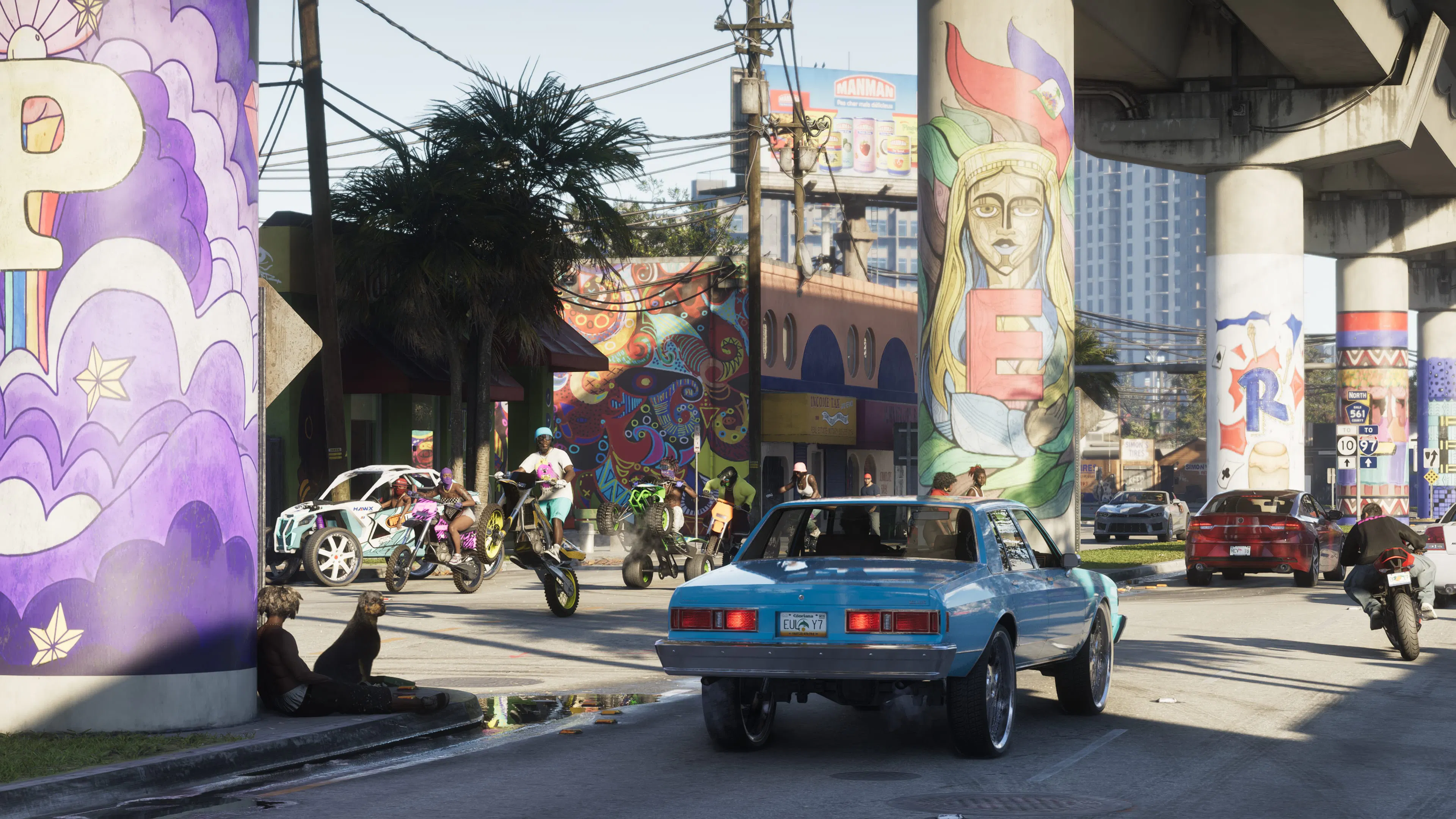 A blue lowrider and dirt bikes under a highway overpass covered in murals.