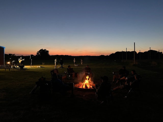 Outside, dusk on a summer day on Long Island, NY, some soldiers from 1-172 Cav reminisce around a fire while others play cornhole and Brian Ripps plays live music in the background. 