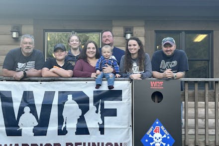 The Smartt family poses for a photo outside of the main lodge at Camp Rotary behind a WRF banner and a 1st Recon Battalion commemorative cornhole board. 