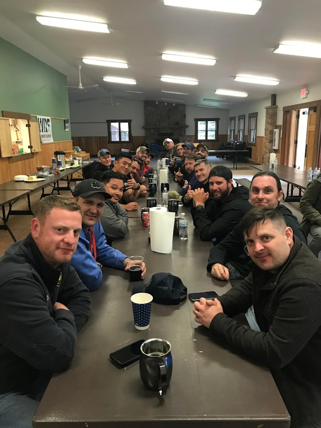 Inside the dining facility at Camp Rotary, Montana, gathered around a long dining table, 20 soldiers of Creek Co 2-4 casually pose for a group photo.
