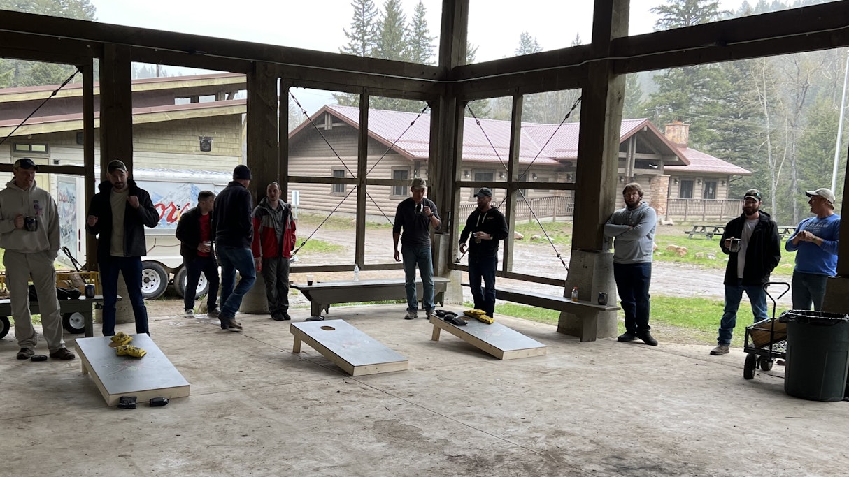 Under a pavilion, on a cloudy day, soldiers of Creek Co 2-4 observe and participate in a cornhole tournament to win the custom boards they are using.