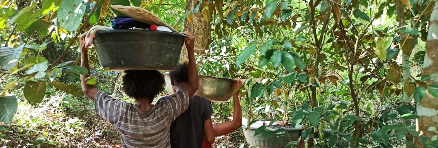 Two people carrying baskets on their heads, walking through forest.