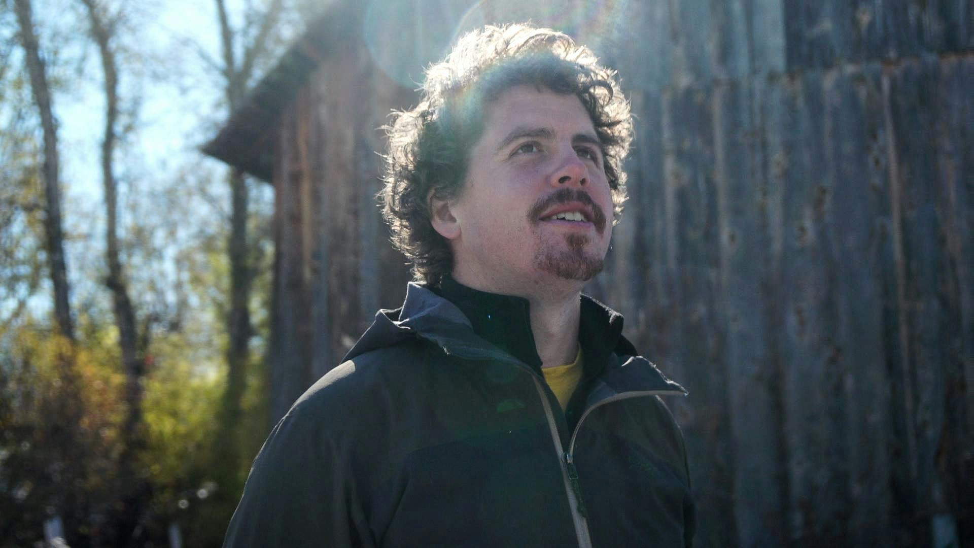 Headshot of Zachary Weiss looking up to the sky illuminated by a ray of sunlight and standing in front of a rustic barn