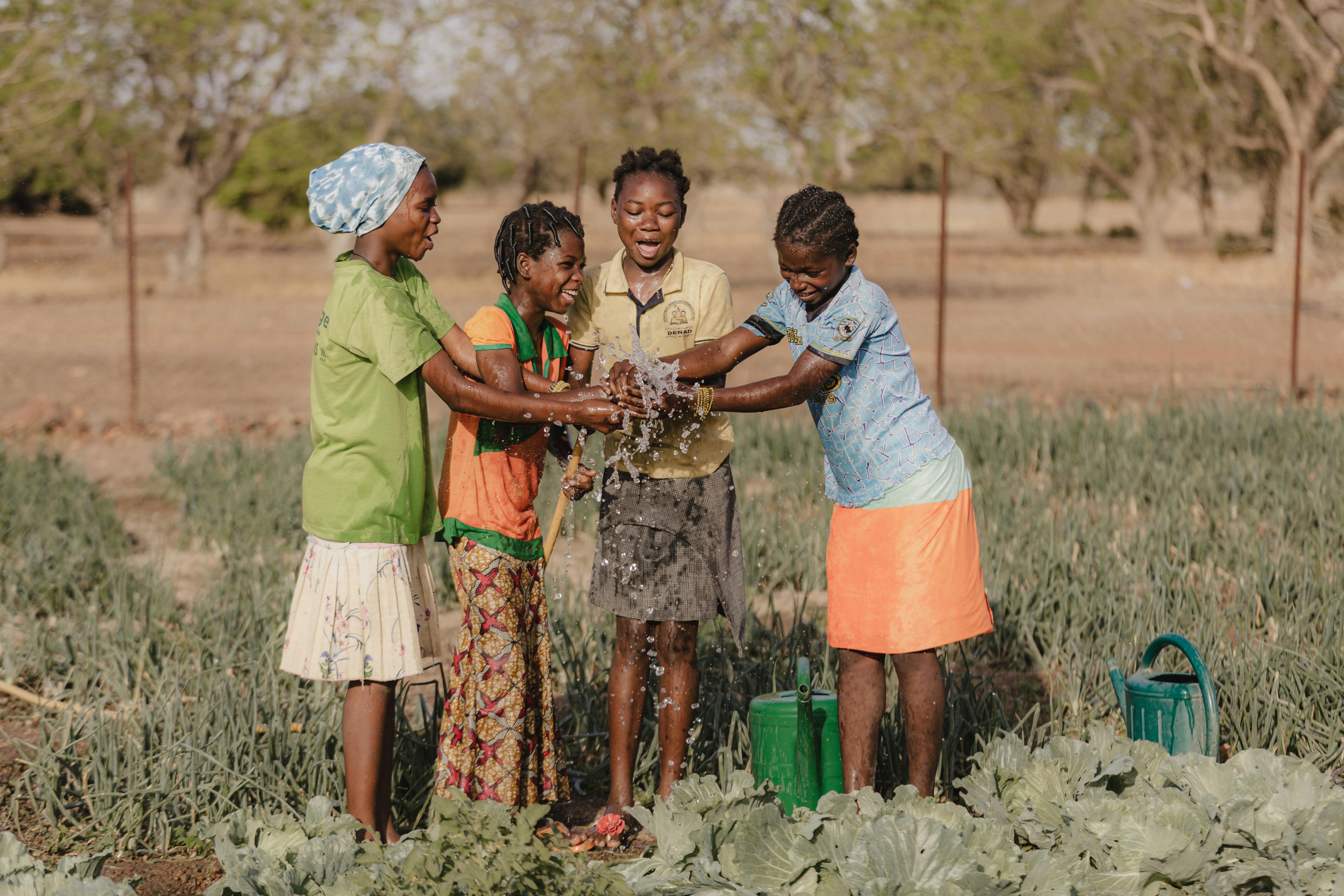 Four children standing in a vegetable garden, holding a stream of water together with green watering cans nearby.