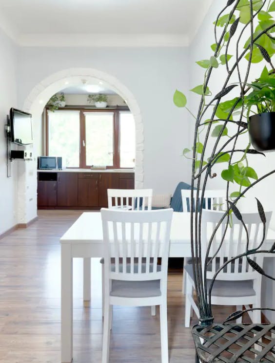 In the image - a bright kitchen with a dining area in white. In the foreground there is a table with chairs, in the background there is a work surface with a view out the window