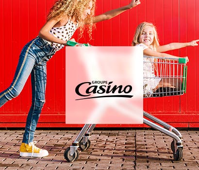 Une mère de famille pousse un caddie avec sa fille dedans, logo du Groupe Casino