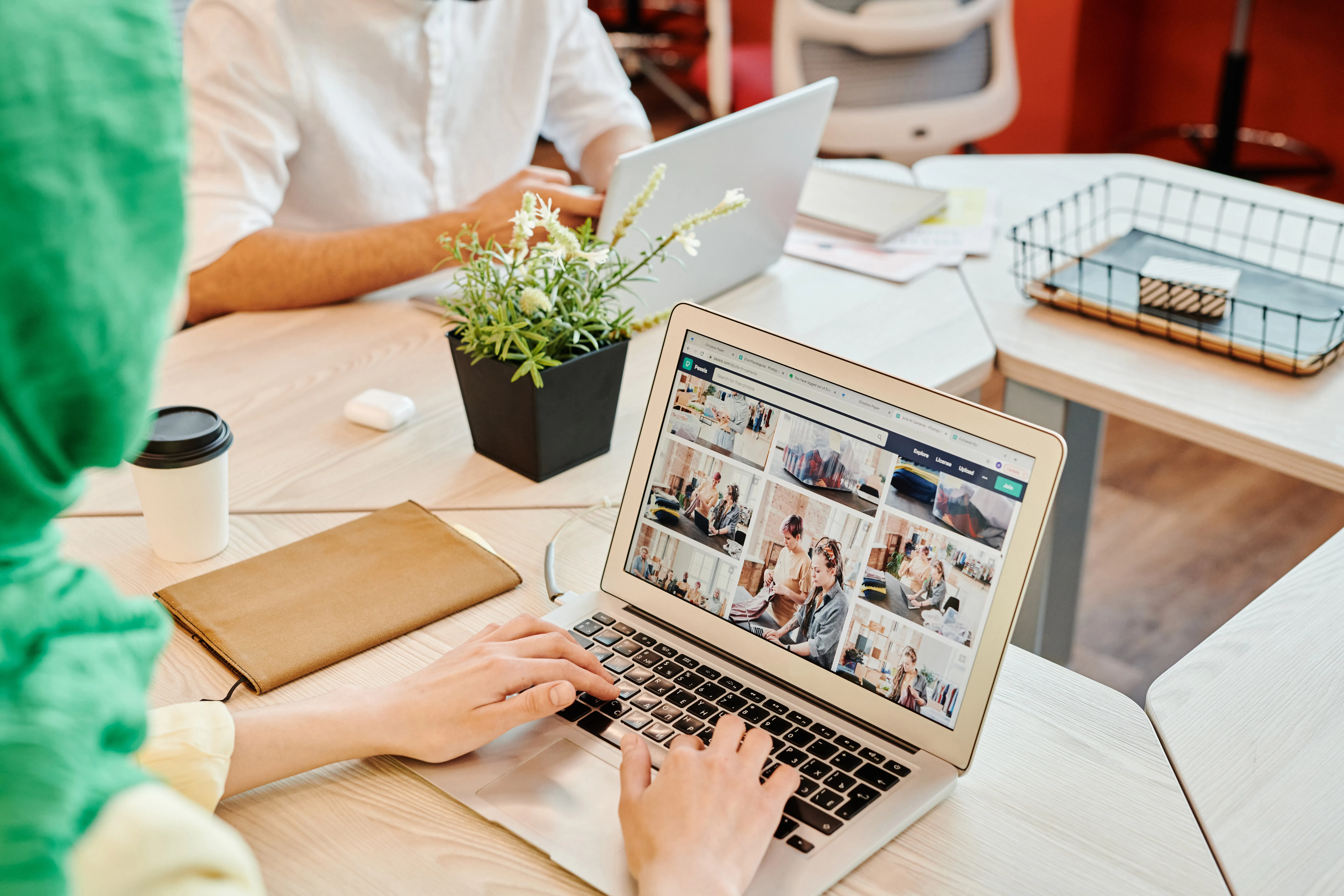Office photo of a person using a laptop showing a grid of images, with coworkers and desk items in the background