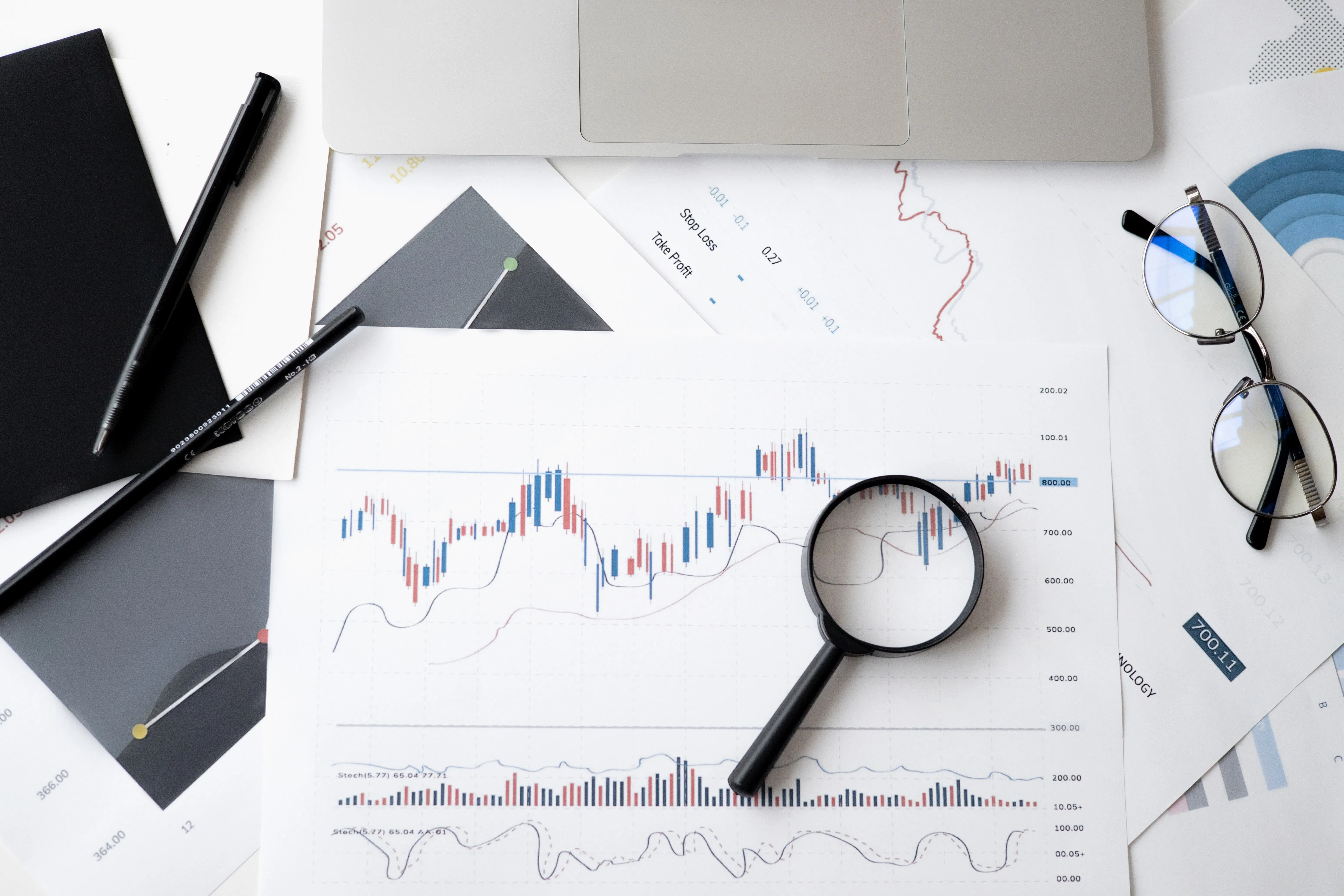 Top-down photo of printed financial charts with a magnifying glass, notebook, and glasses on a desk