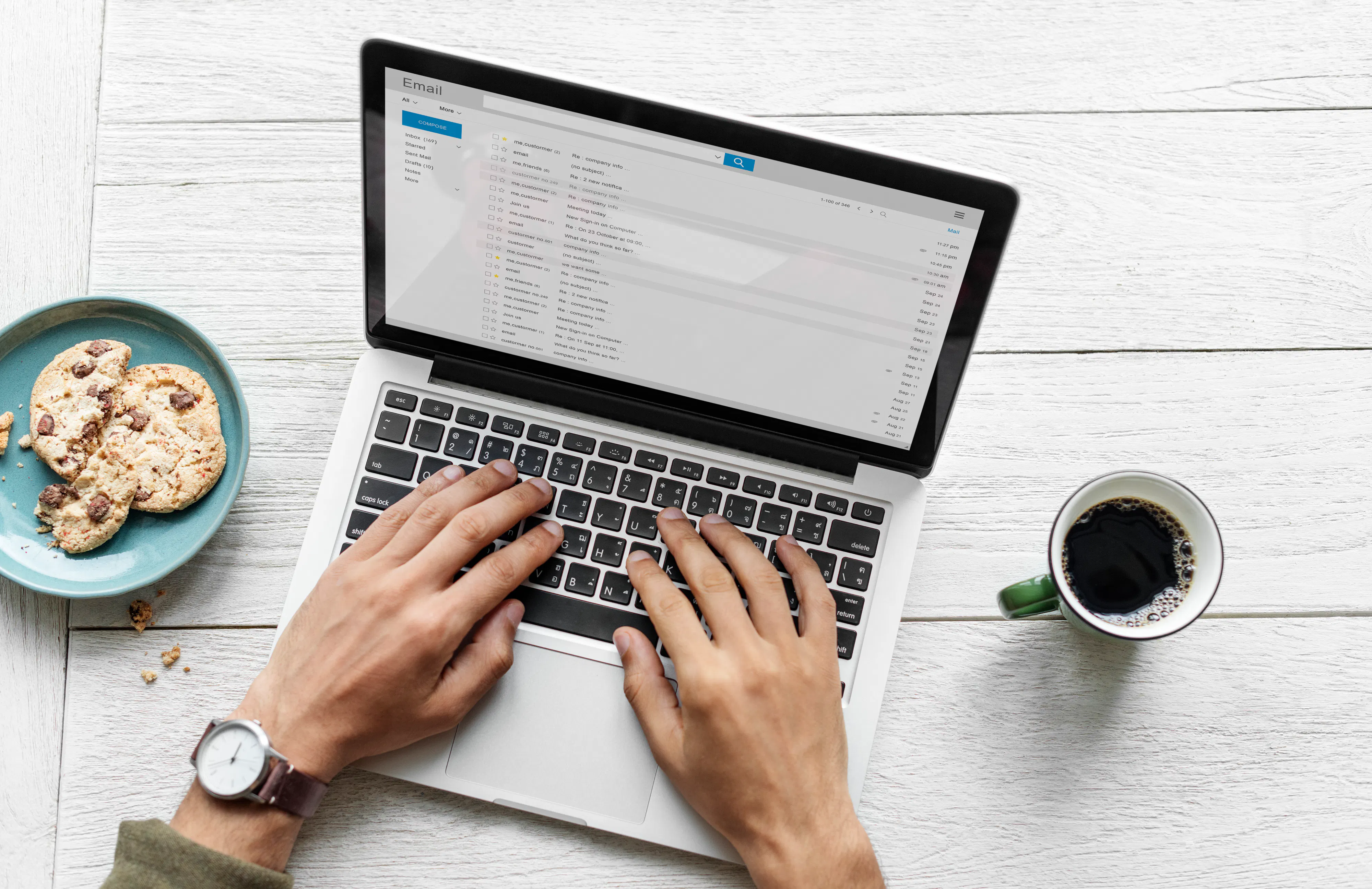 Hands typing on a laptop on a desk with coffee and cookies on a plate nearby