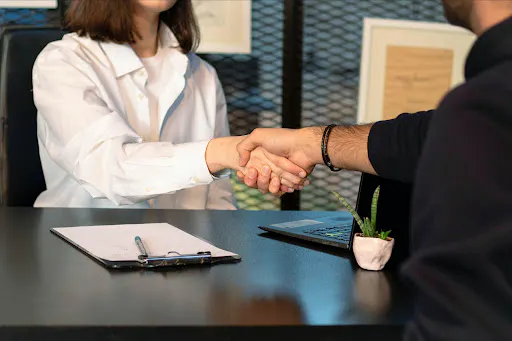 Two people shaking hands across a desk with paperwork, suggesting a job offer or interview agreement