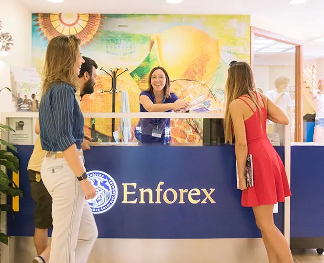 Reception Enforex con due studenti che parlano con una receptionist in un ambiente luminoso e moderno.
