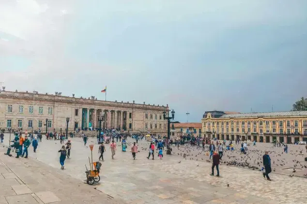 Praça em frente ao Parlamento em Lisboa, com grande área pavimentada, pessoas caminhando e bandeira ao fundo sob céu nublado, cenário turístico da capital portuguesa.