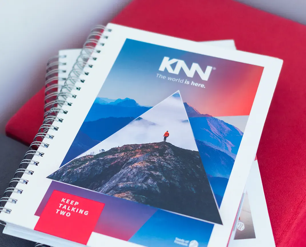 Caderno espiral com capa e páginas de design gráfico, exibindo a marca KNN e imagens de paisagens de montanhas, céu e pôr do sol em tons azul e vermelho.