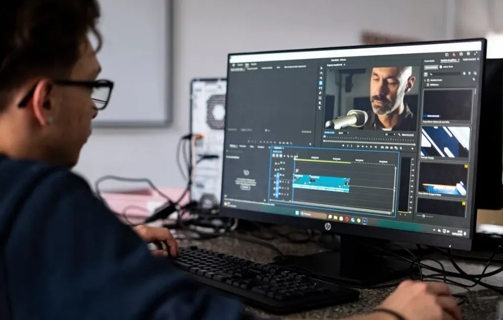Homem trabalhando em computador com softwares de edição e comunicação, analisando uma interface de áudio e vídeo em múltiplas janelas, em ambiente de estúdio.