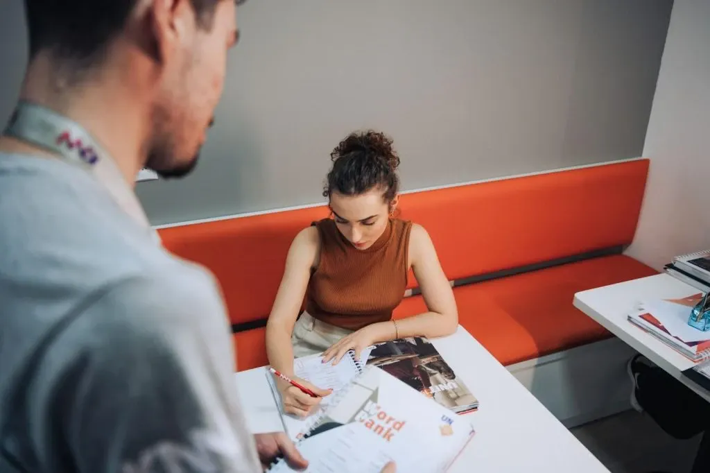 Jovem sentada em uma área de escritório com bancos laranja, folheando revistas ou materiais impressos enquanto um homem ao lado observa, em ambiente de trabalho colaborativo.