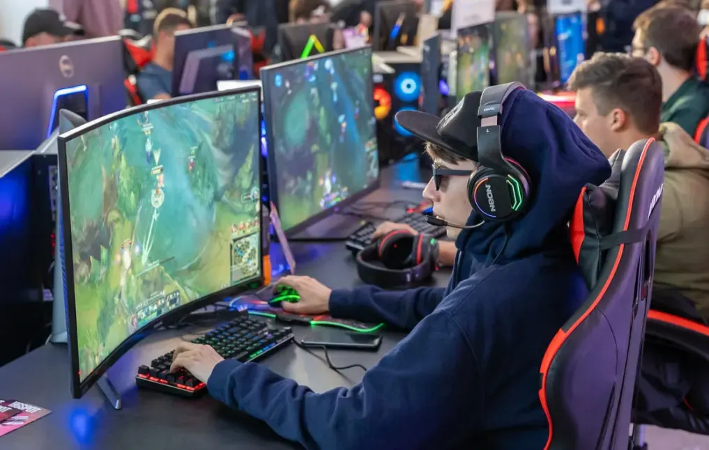Jogador em campeonato de games usando headset e mouse, com computadores monitorando um jogo de ação em equipe durante o torneio