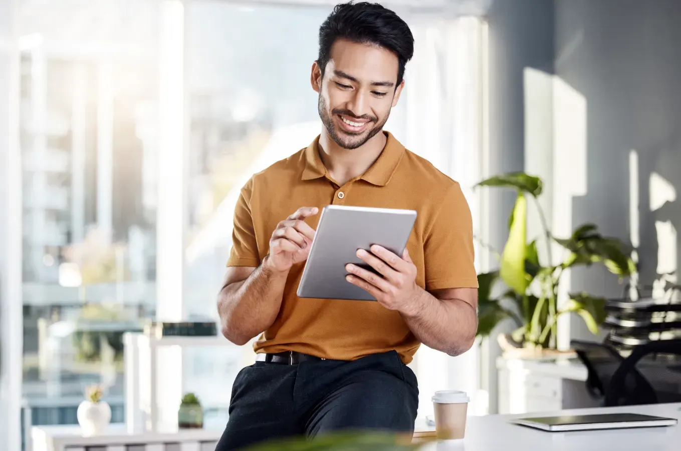 Jovem profissional de camisa polo marrom usando um tablet enquanto sorri em ambiente de trabalho moderno, com escritório ao fundo e copo de café sobre a mesa.