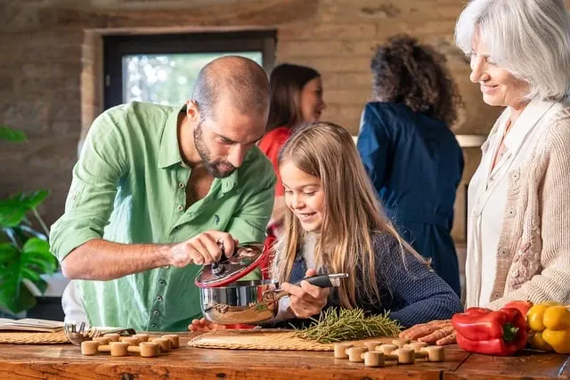 Família reunida na cozinha: pai prepara uma receita com a filha pequena, enquanto a avó e outras pessoas ao fundo observam. Ingredientes e massa sobre a bancada.