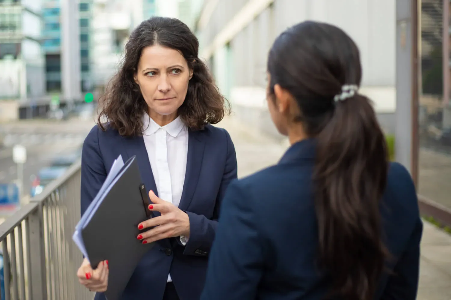 Mulher executiva em reunião de negócios ao ar livre, conversando com outra pessoa ao lado de um escritório, segurando uma pasta e olhando atentamente durante a conversa.