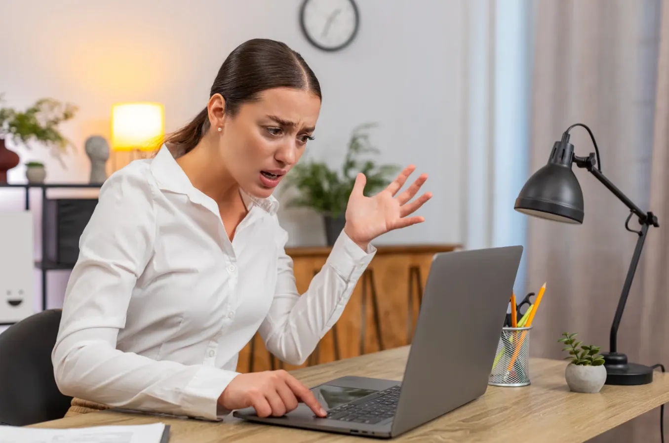 Mulher em home office, usando camisa branca e trabalhando no notebook, expressa surpresa ou preocupação enquanto parece dar instruções com a mão no escritório moderno com luminária e planta ao fundo.
