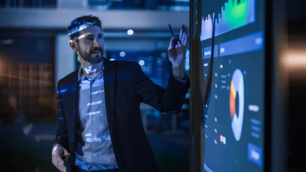 Homem em sala de controle, com tela e gráficos de dados no fundo, usando um dispositivo e tocando a tela digital com gráficos de mercado em tempo real, estilo tecnologia e finanças.