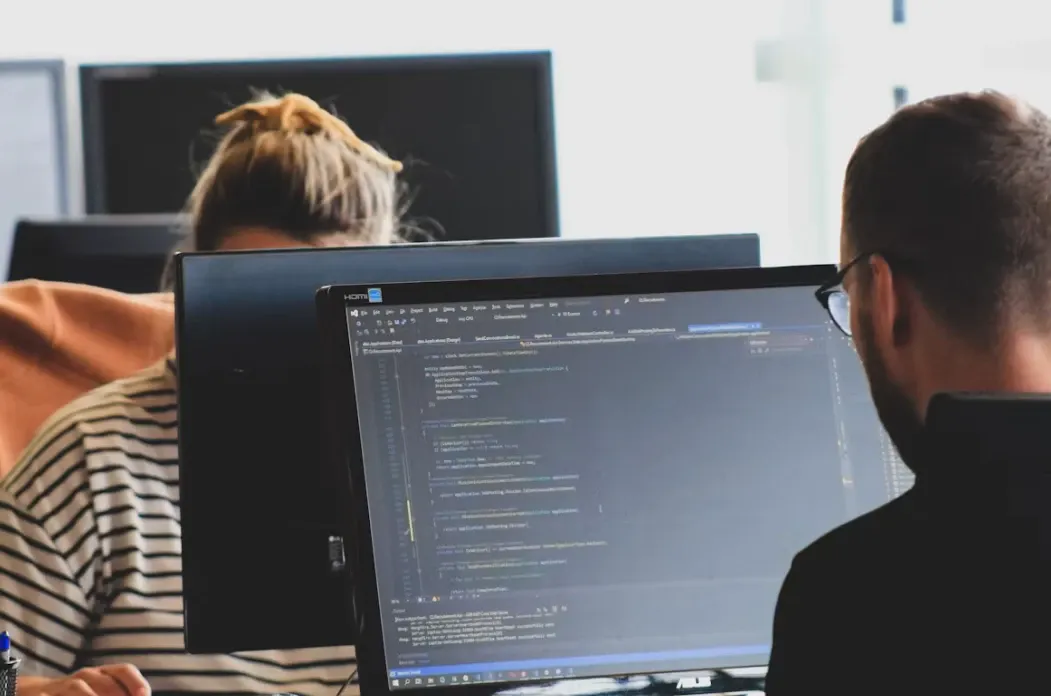 Equipe trabalhando em computadores com código de programação visível em uma tela, escritório com desenvolvedor olhando para o monitor e outro profissional ao fundo, foco em tecnologia e desenvolvimento de software.