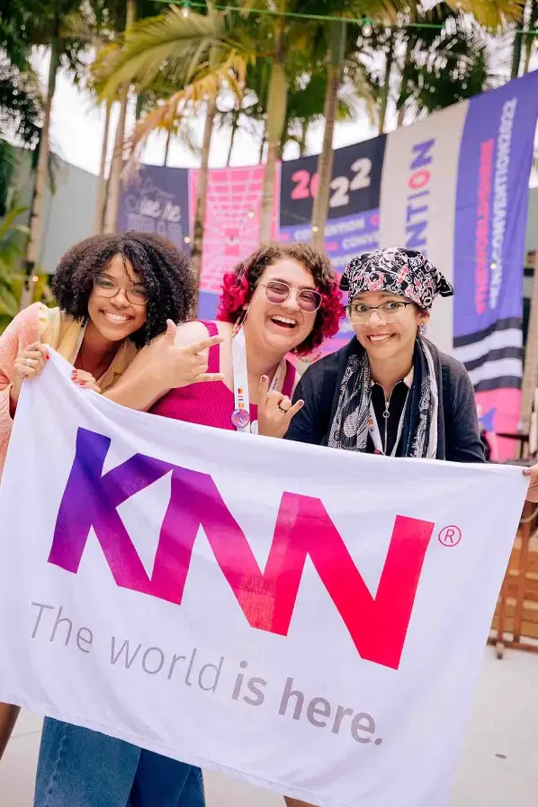 Três participantes sorridentes em evento ao ar livre seguram uma bandeira grande da marca KNN, com palmeiras ao fundo e painéis coloridos ao estilo de convenção.