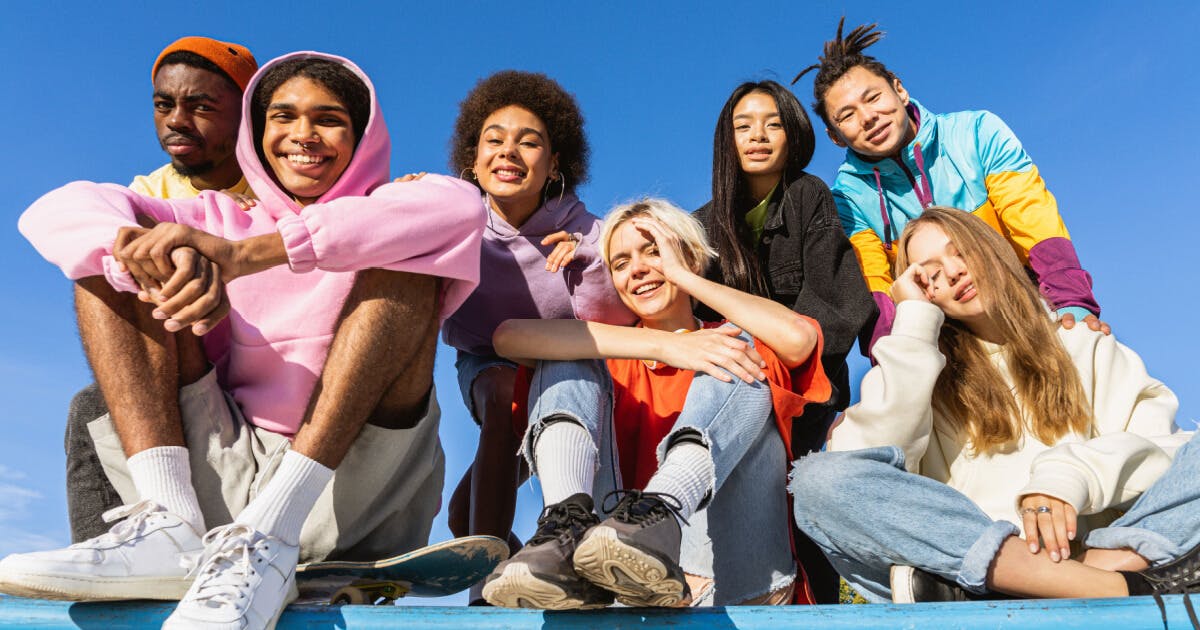 Grupo de jovens sentados em um skate, sorrindo e se divertindo em um ambiente ao ar livre.