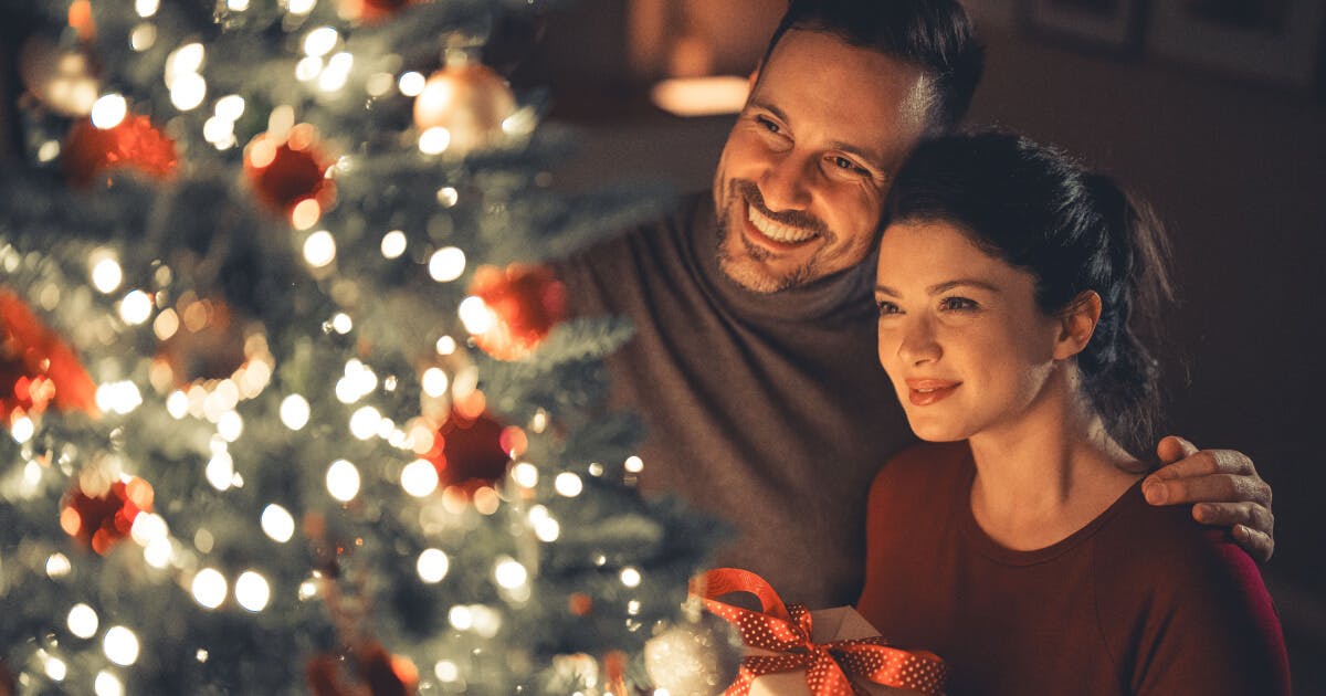 Um homem e uma mulher sorriem diante de uma árvore de Natal decorada.